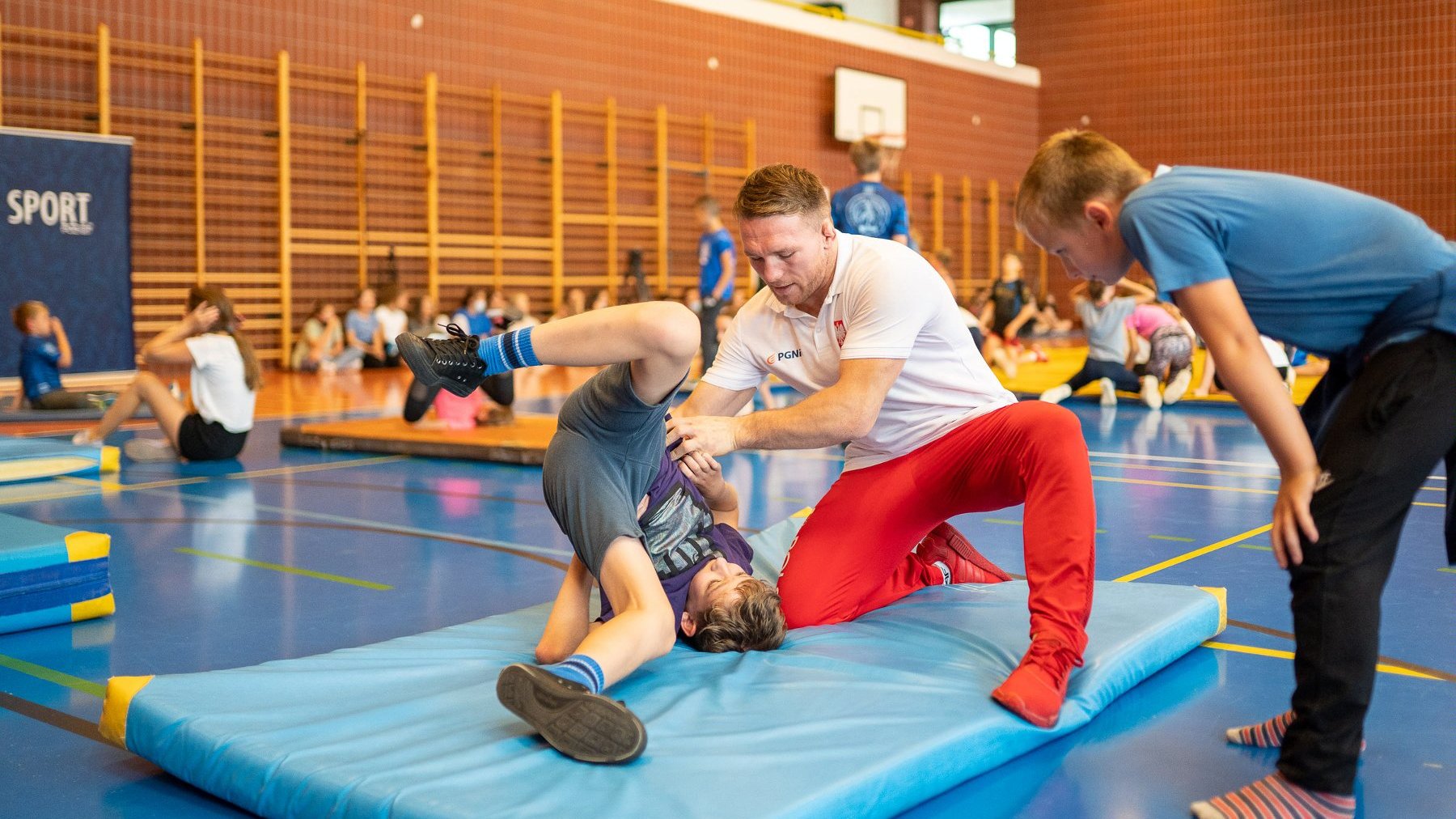 Na zdjęciu sala gimnastyczna i medalista olimpijski trenujący ucznia na macie, obok inne dzieci