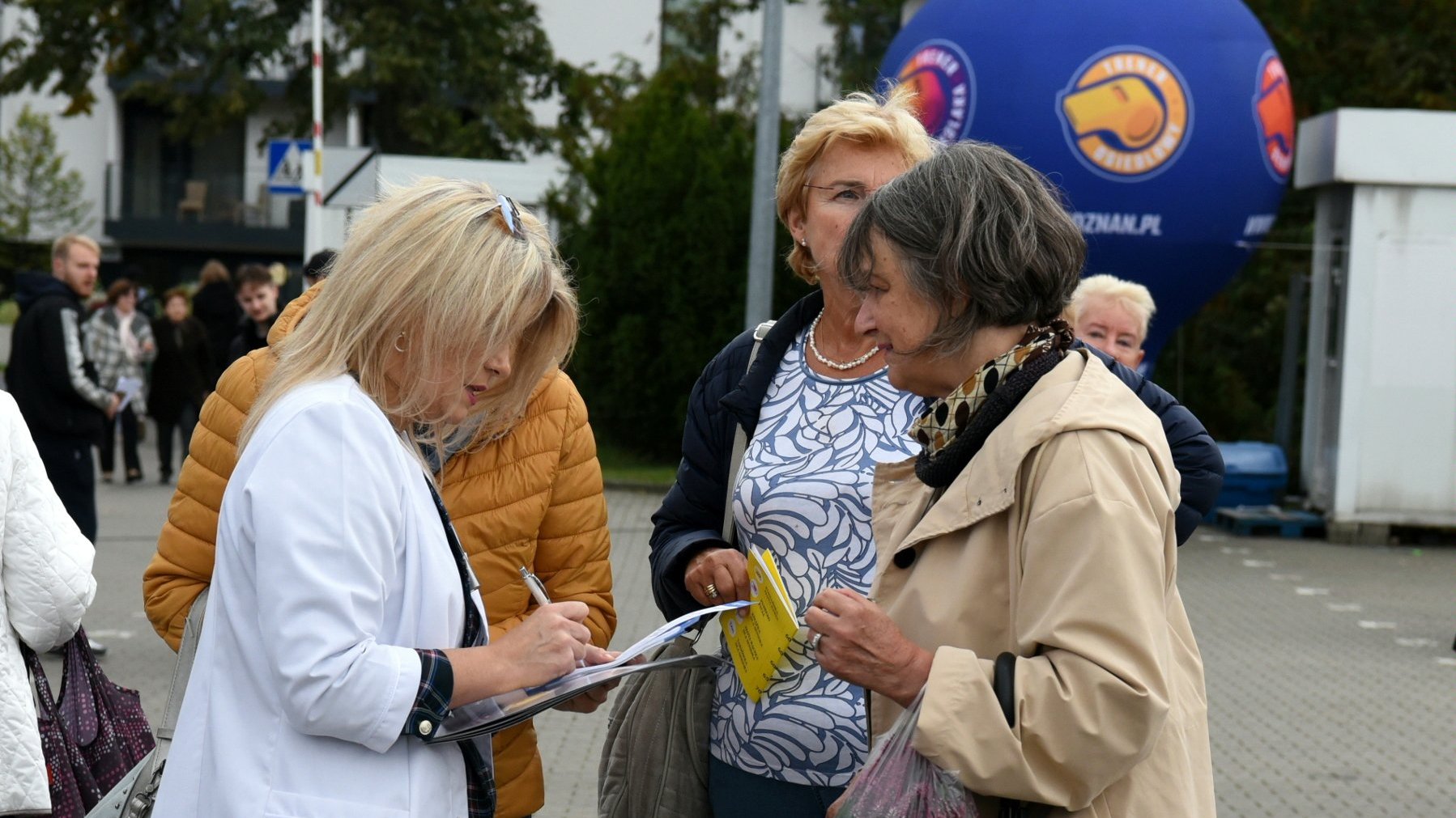 Kobieta w medycznym fartuchu, z długopisem w ręku, tłumaczy coś trzem seniorkom