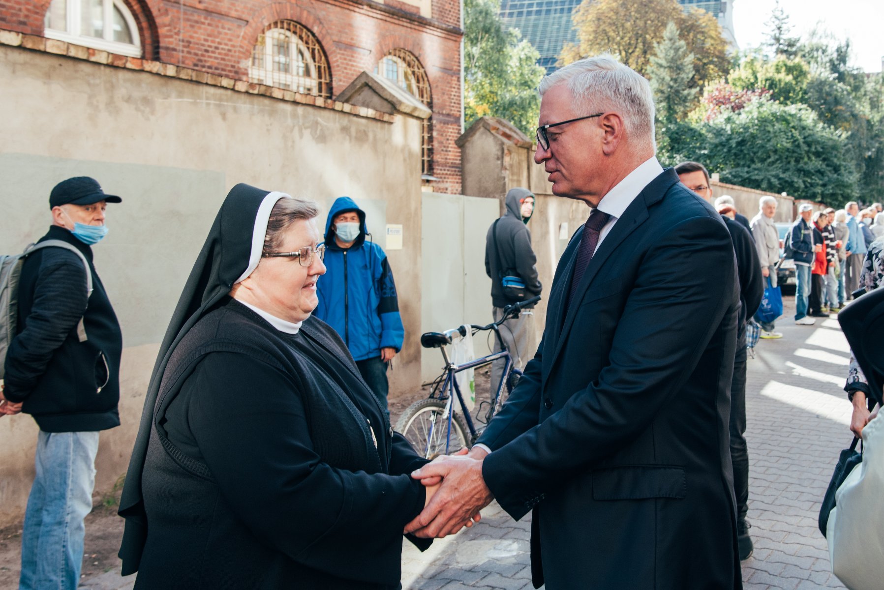 Na zdjęciu Jacek Jaśkowiak, prezydent Poznania, ściskający dłonie siostry Józefy, w tle kolejka ludzi - grafika rozmowy