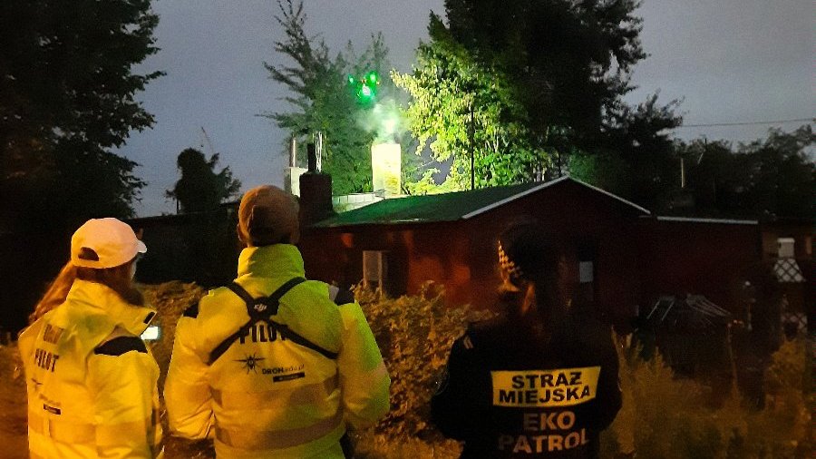 Zdjęcie przedstawia latającego drona oraz pracowników, którzy go obsługują. Fotografię zrobiono po zmroku.