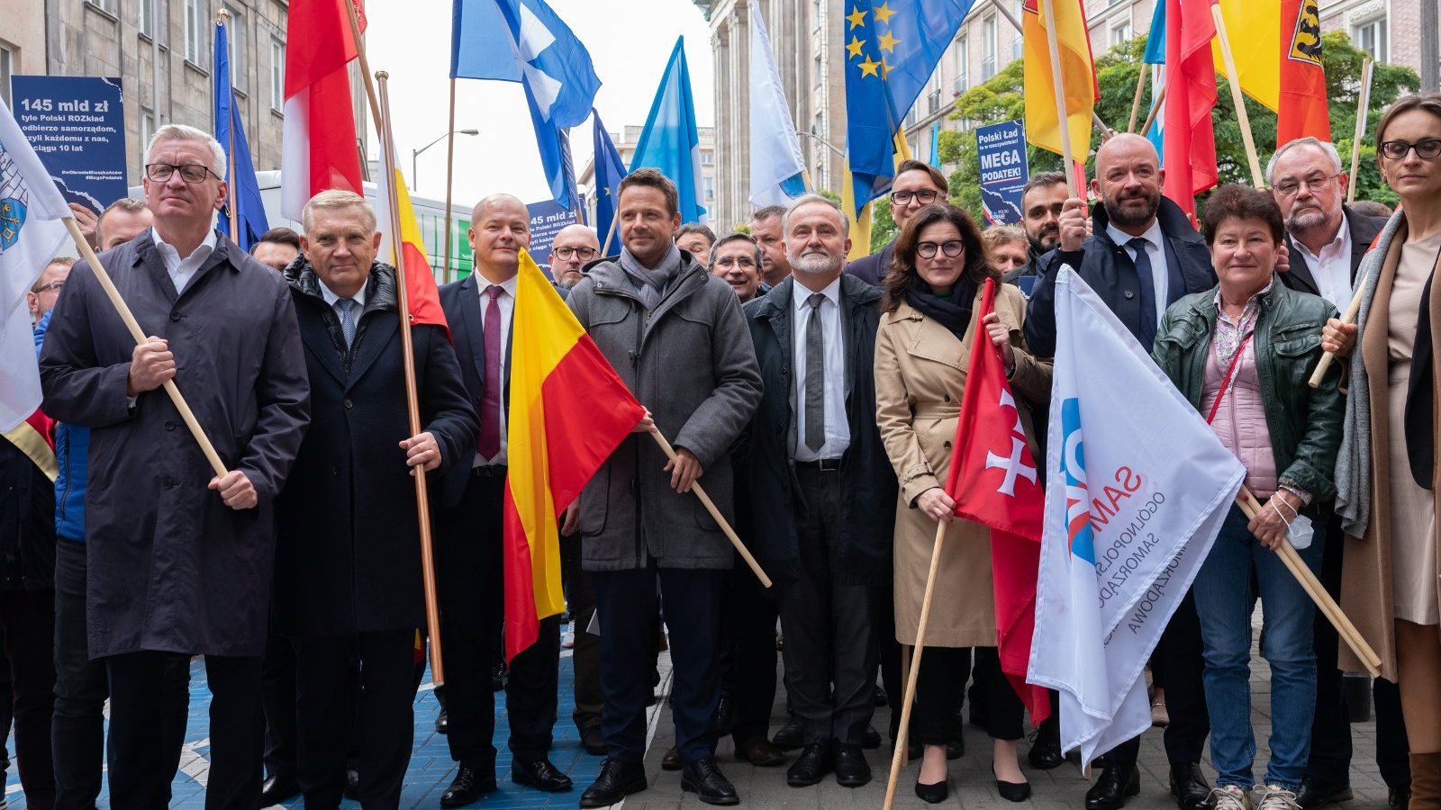 Samorządowcy stojący obok siebie, trzymający kolorowe flagi swoich z herbami swoich miejscowości