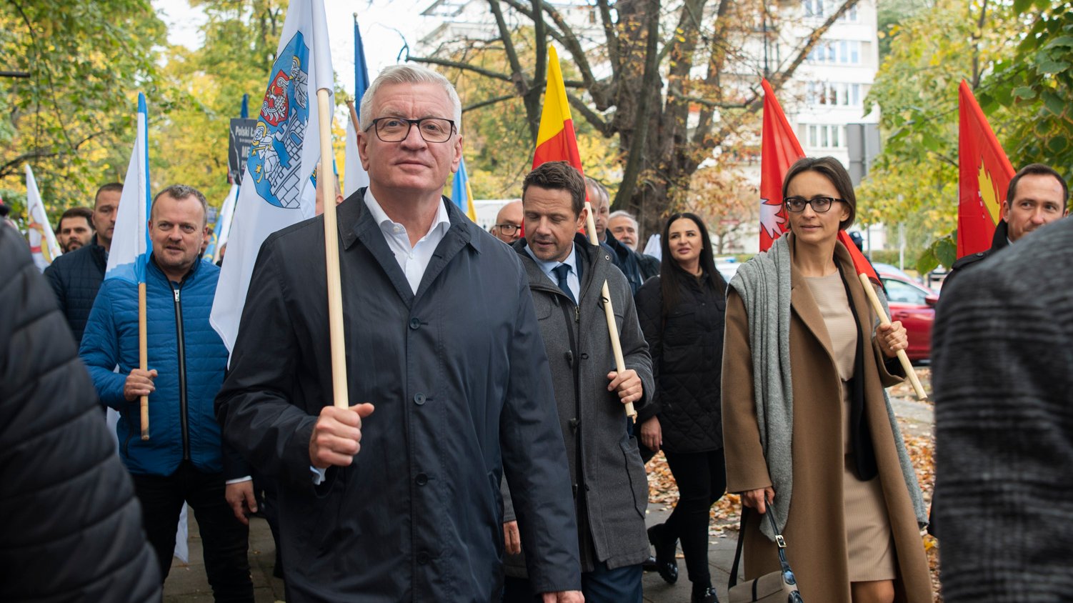 Na zdjęciu samorządowcy z flagami swoich miejscowości, idą ulicą, na pierwszym planie Jacek Jaśkowiak, prezydent Poznania