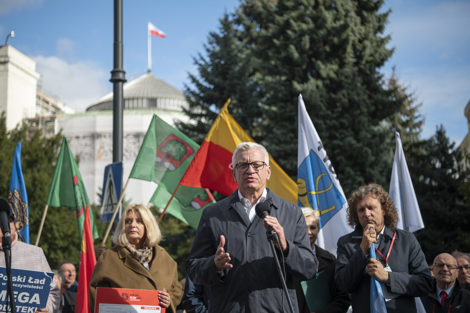 Na zdjęciu Jacek Jaskowiak, prezydent Poznania, przy mikrofonie, za nim inni samorządowcy z flagami, w tle budynek Sejmu - grafika rozmowy