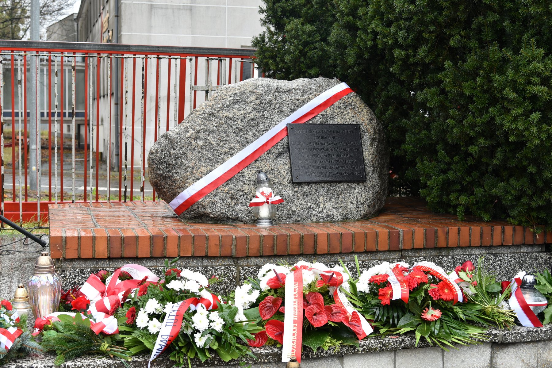 Zdjęcie przedstawia głaz upamiętniający miejsce, gdzie od końca października 1939 r. do maja 1940 r. działał niemiecki obóz przesiedleńczy. Jest on przepasany biało czerwoną wstęgą, pod nim znajdują się kwiaty. - grafika rozmowy