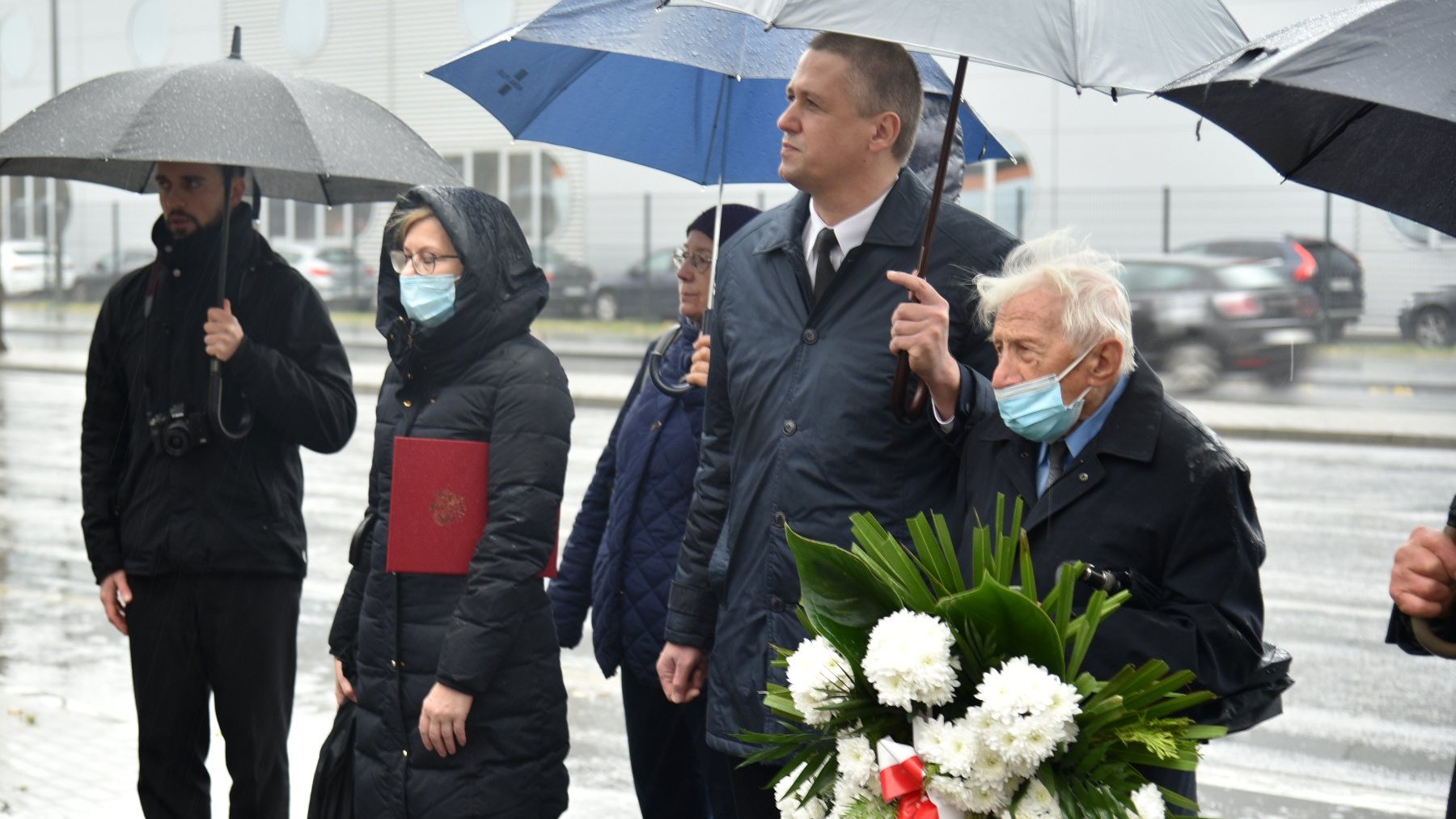 Zdjęcie przedstawia stojących ludzi pod parasolami. Jeden z mężczyzn trzyma kwiaty.