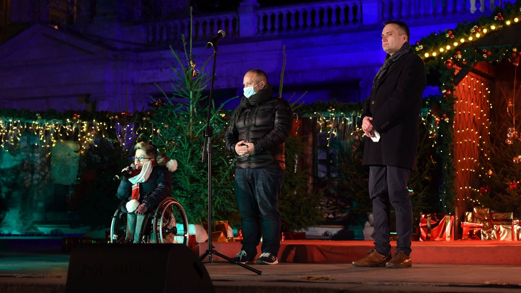 Na zdjęciu trzy osoby na scenie, dwaj mężczyźni stoją, kobieta siedzi na wózku, w tle choinki