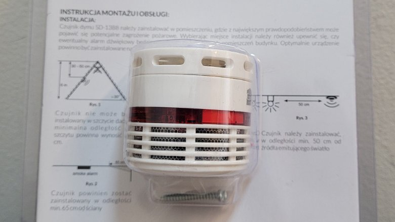 Na zdjęciu czujnik dymu - niewielkie białe urządzenie, zapakowane w przezroczyste opakowanie