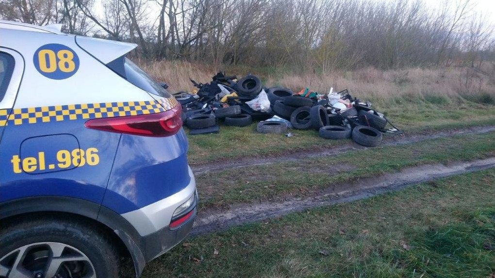 Hałda starych opon i innych śmieci leżąca na trawie. Z lewej strony kadru fragment samochodu straży miejskiej z wiidoczmym numerem alaromowym - 986.