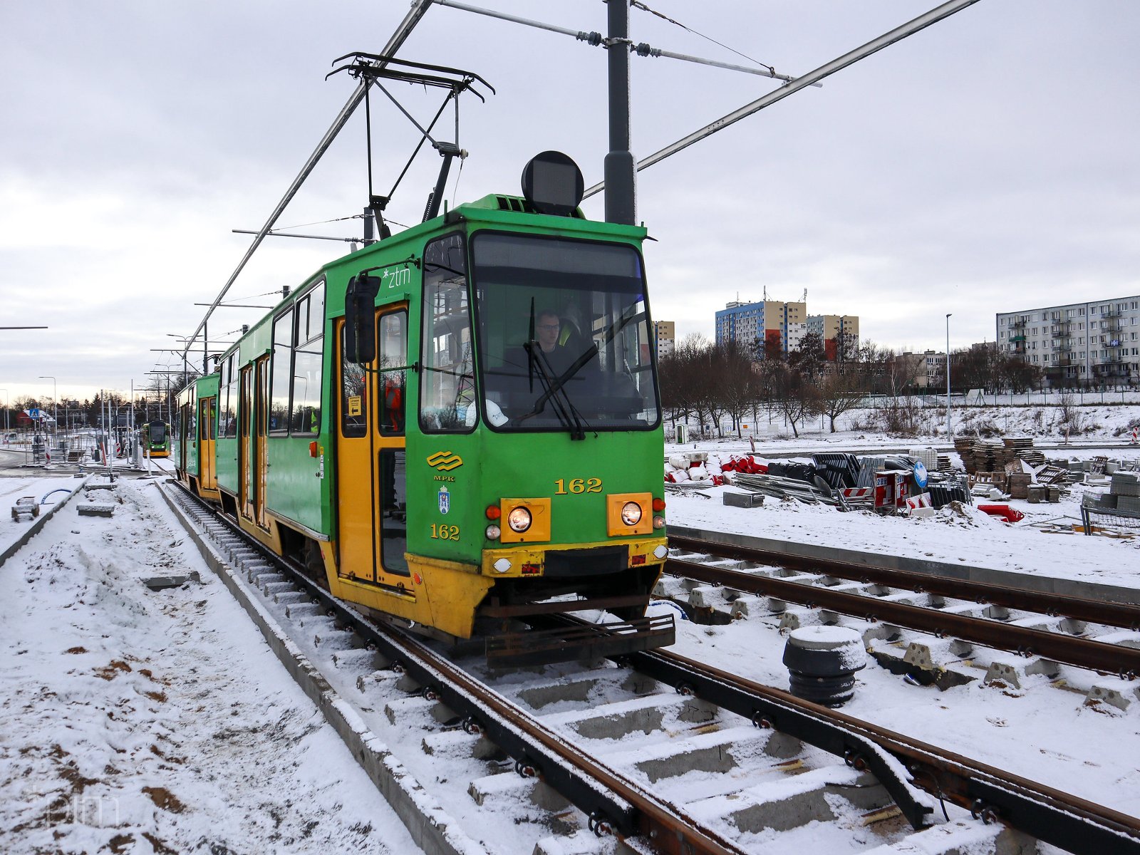 Tramwaj stojący na torowisku, w tle bloki, na trawie śnieg - grafika rozmowy