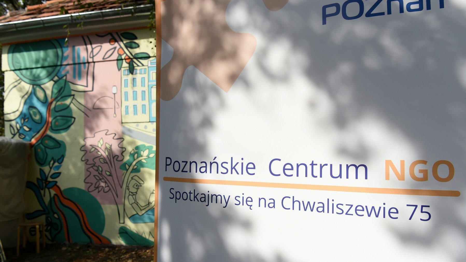 Na zdjęciu podwórko, na pierwszym planie baner z nazwą Centrum, w tle kolorowa ściana z muralem - grafika rozmowy