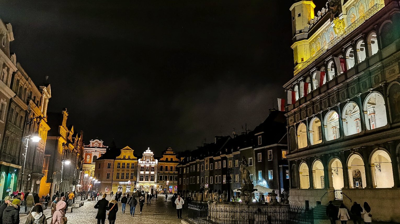 Zdjęcie przedstawia Stary Rynek po zmroku. Widać na nim budynki i spacerujących ludzi. - grafika rozmowy