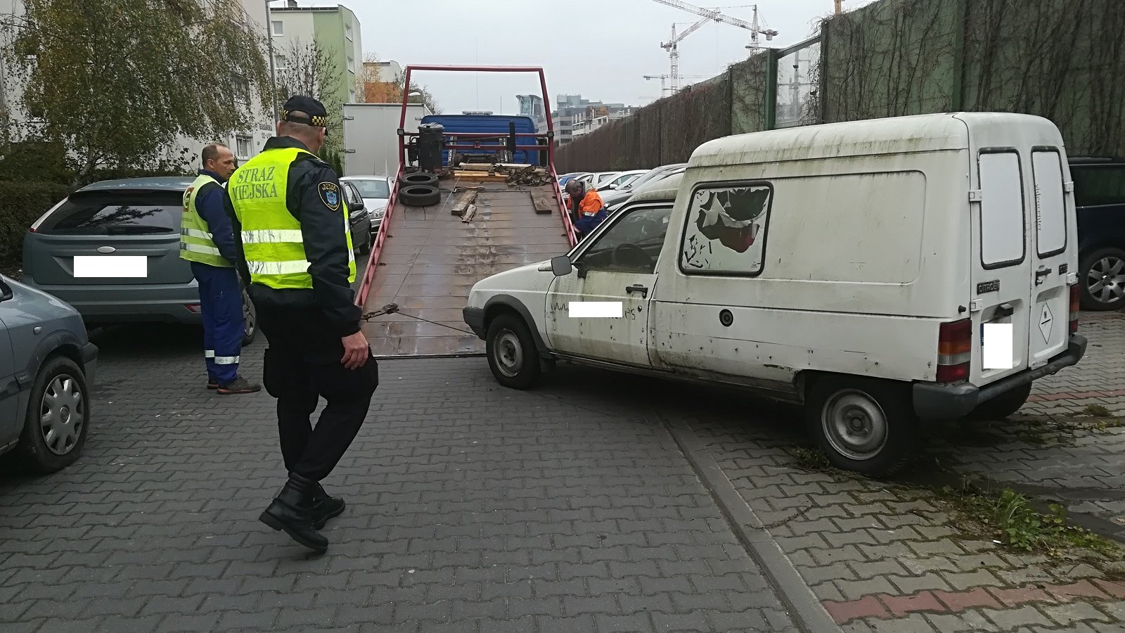 Zdjęcie przedstawia strażników miejskich, którzy nadzorują akcję wywozu wraków. Widać na nim dwóch mężczyzn, lawetę i kilka stojących aut.