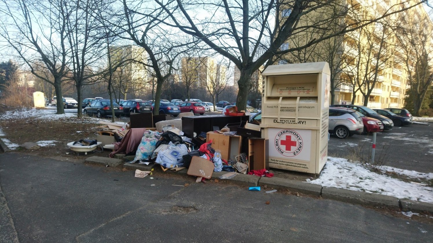 Zdjęcie przedstawia kontener PCK na ubrania, stojący na osiedlu wśród bloków i aut. Obok znajduje się hałda różnego rodzaju śmieci.