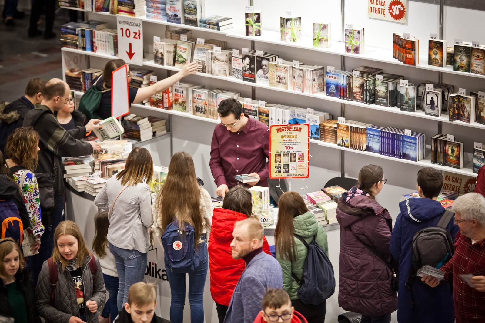 Zdjęcie przedstawia dużą półkę z książkami i ladę, przed którą stoi tłum ludzi. - grafika rozmowy