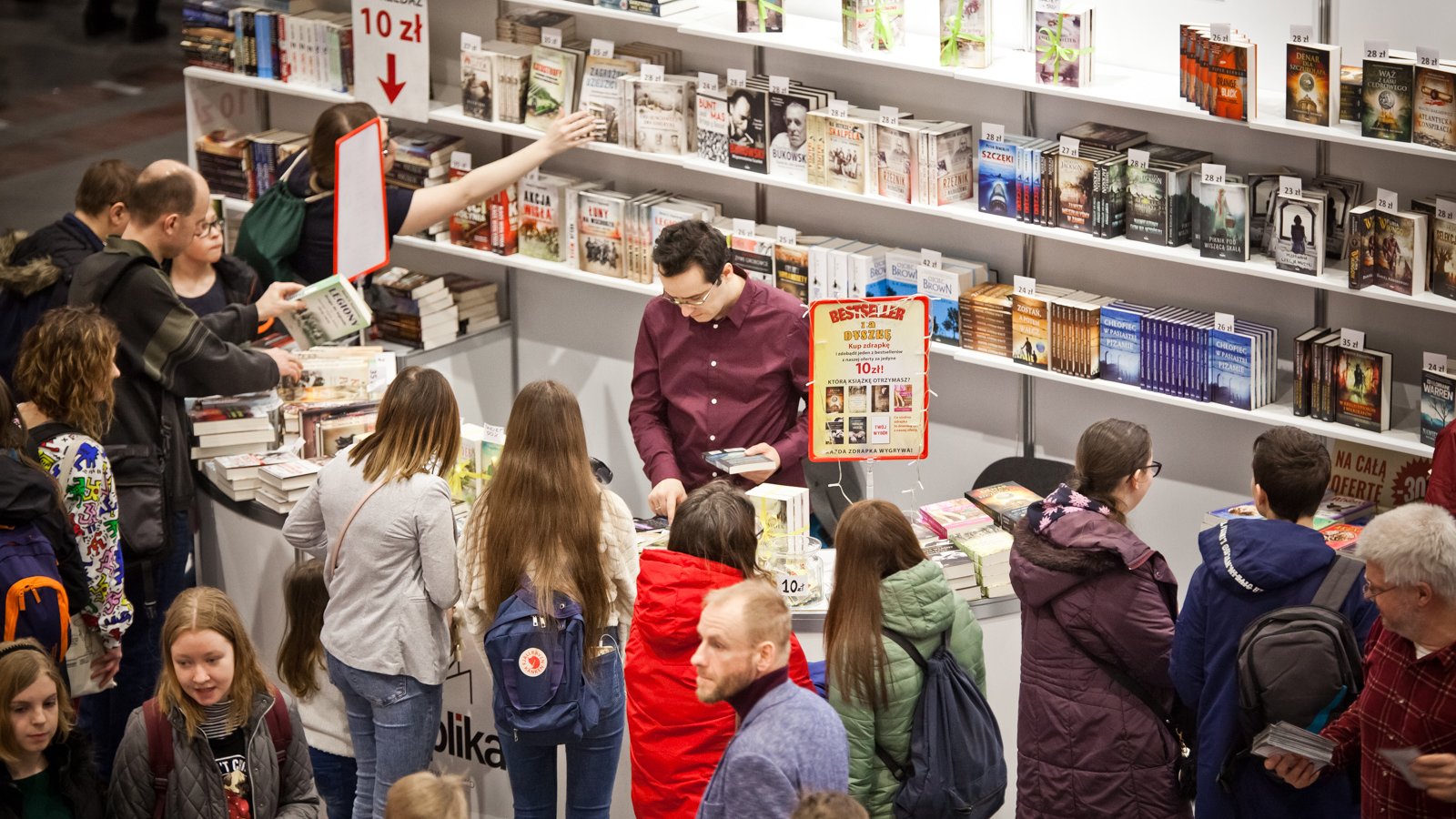 Zdjęcie przedstawia dużą półkę z książkami i ladę, przed którą stoi tłum ludzi.