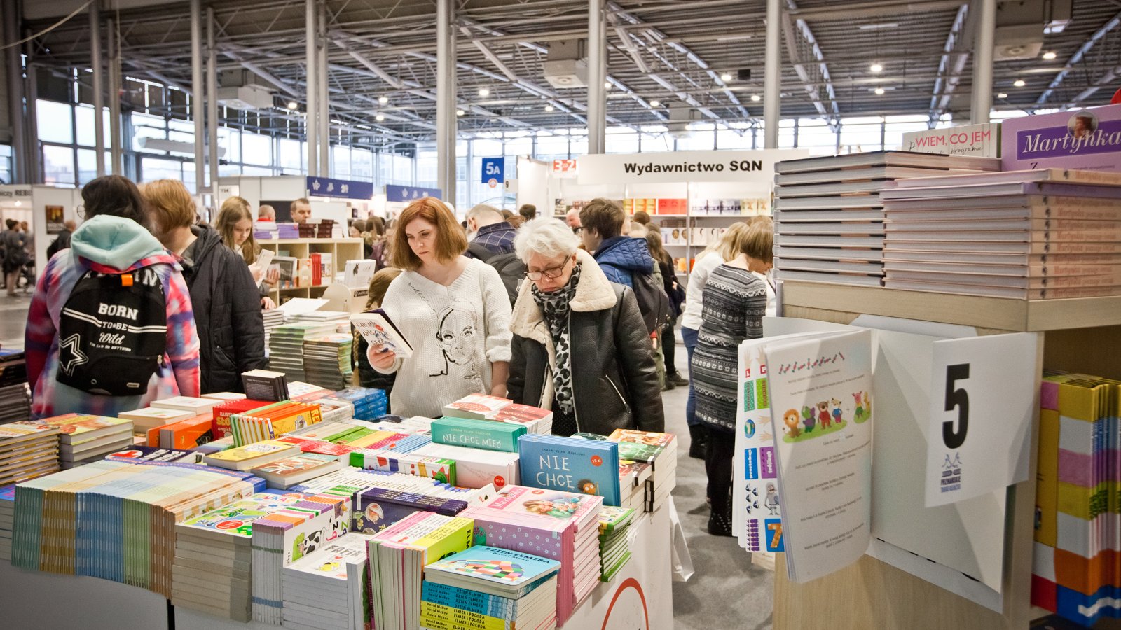 Zdjęcie przedstawia ludzi oglądających książki oraz dużo stoisk z książkami.