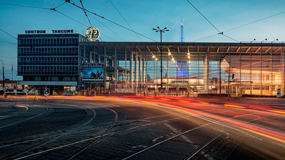 Na zdjęciu wejście główne na Międzynarodowe Targi Poznańskie, na pierwszym planie ślady świateł samochodów - grafika rozmowy