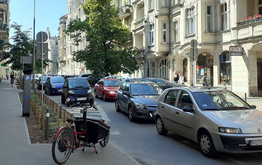 Na zdjęciu poznańska ulica, na niej samochody, na pierwszym planie chodnik i zaparkowany na nim rower - grafika rozmowy