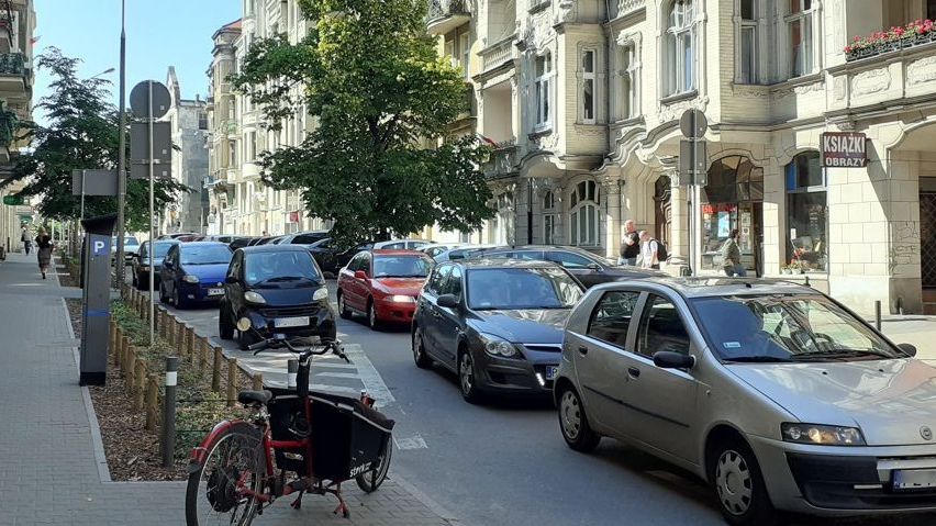 Na zdjęciu poznańska ulica, na niej samochody, na pierwszym planie chodnik i zaparkowany na nim rower