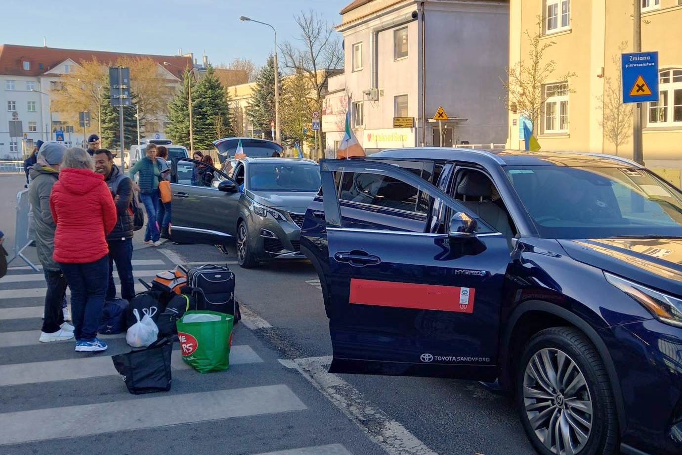 Na zdjęciu grupa ludzi z bagażami wchodzi do rzędu zaparkowanych samochodów - grafika rozmowy