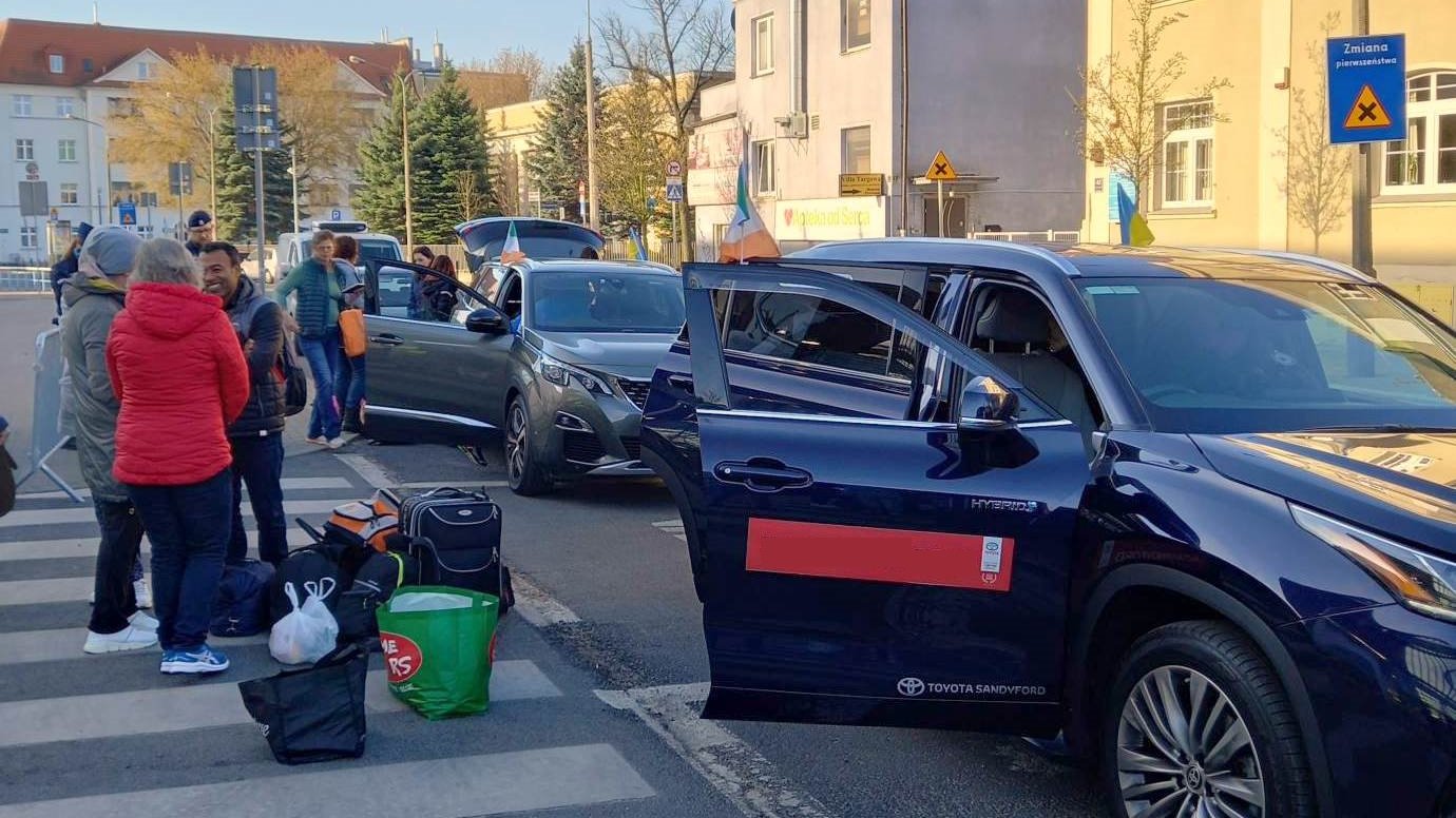 Na zdjęciu grupa ludzi z bagażami wchodzi do rzędu zaparkowanych samochodów