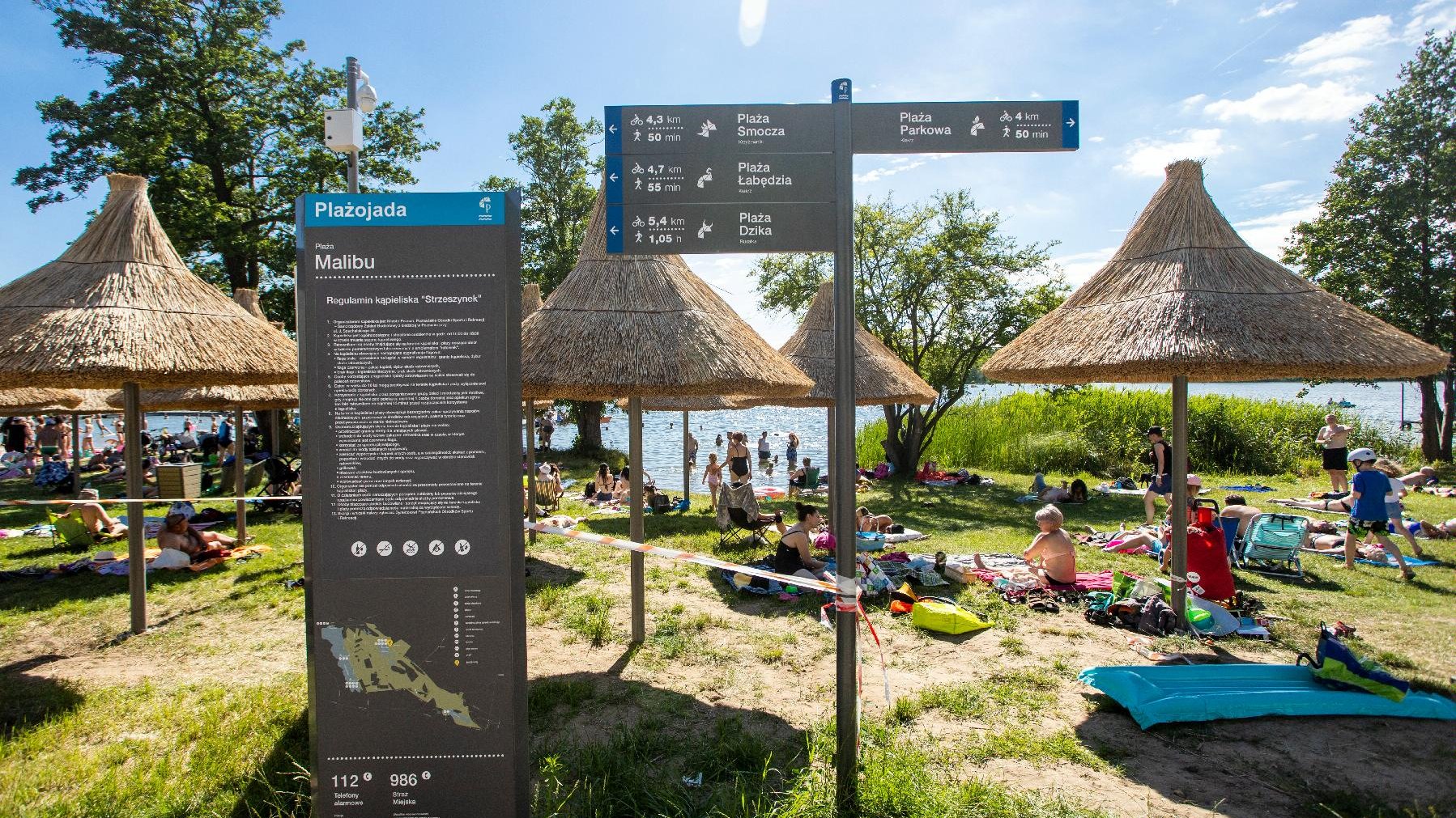 Zdjęcie przedstawia ludzi wypoczywających na plaży pod słomkowymi parasolami. W tle widać jezioro. - grafika rozmowy