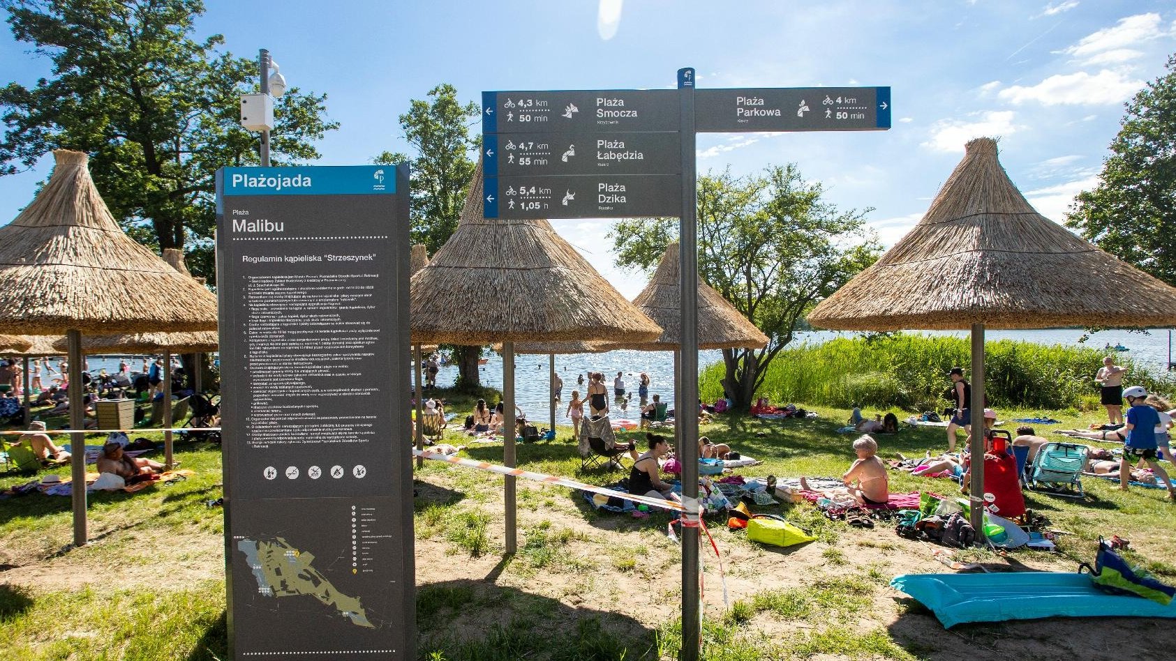 Zdjęcie przedstawia ludzi wypoczywających na plaży pod słomkowymi parasolami. W tle widać jezioro.