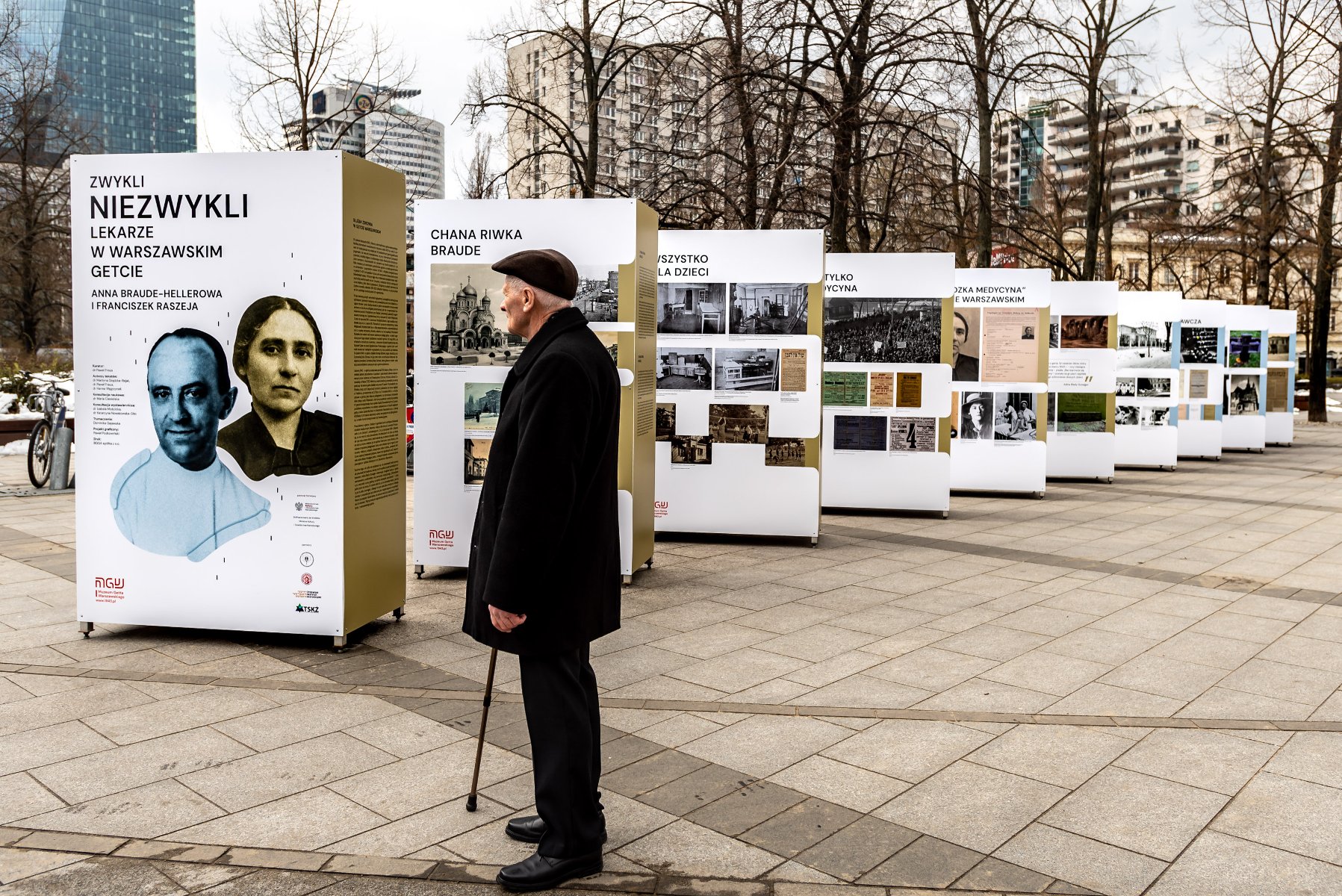 Na zdjęciu wystawa na świeżym powietrzu, kilkanaście stojaków, przygląda im się starszy mężczyzna w ciemnym płaszczu - grafika rozmowy