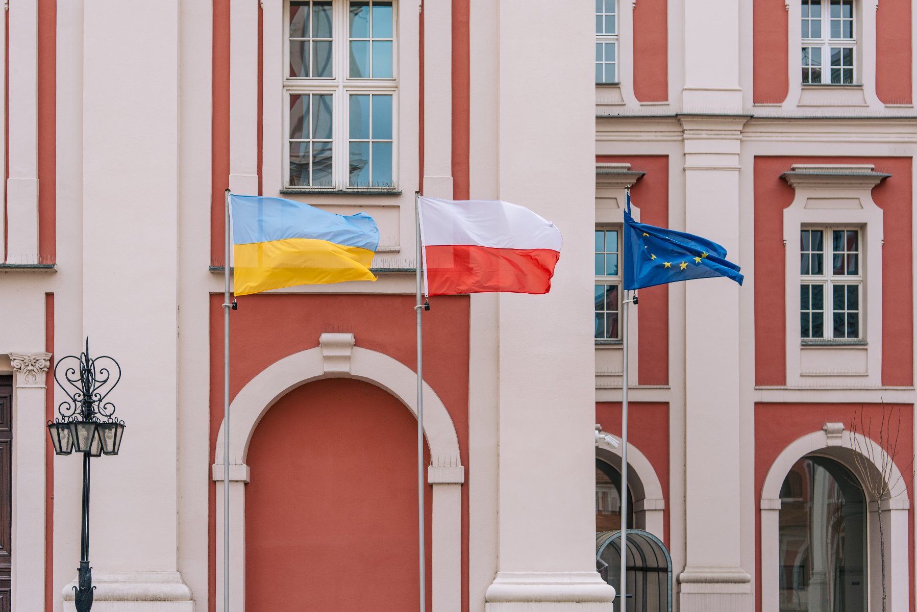 Na zdjęciu widać trzy flagi na tle budynku urzędu miasta. Od lewej: flaga Ukrainy, Polski i Unii Europejskiej. - grafika rozmowy