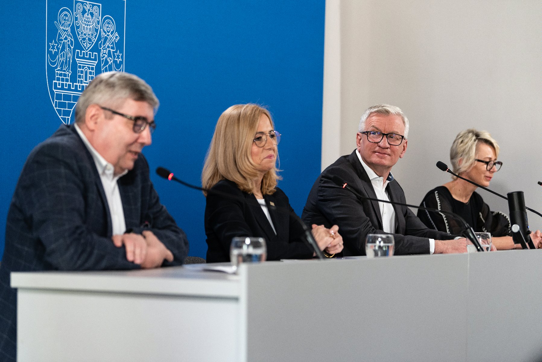 Na zdjęciu cztery osoby za stołem konferencyjnym, w środku prezydent Poznania, w tle granatowa ścianka - grafika rozmowy