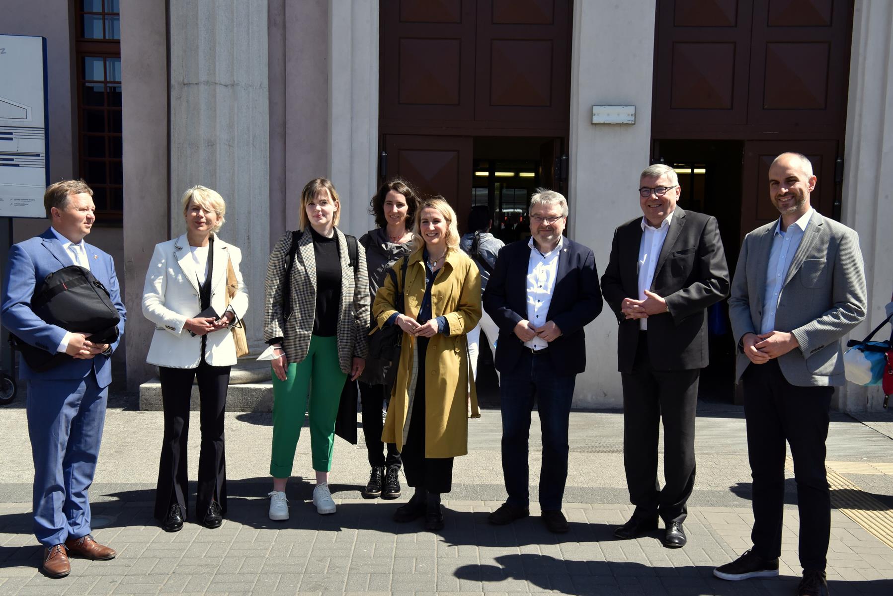 Na zdjęciu delegacja - grupa ludzi stojących obok siebie, za nimi budynek dworca - grafika rozmowy