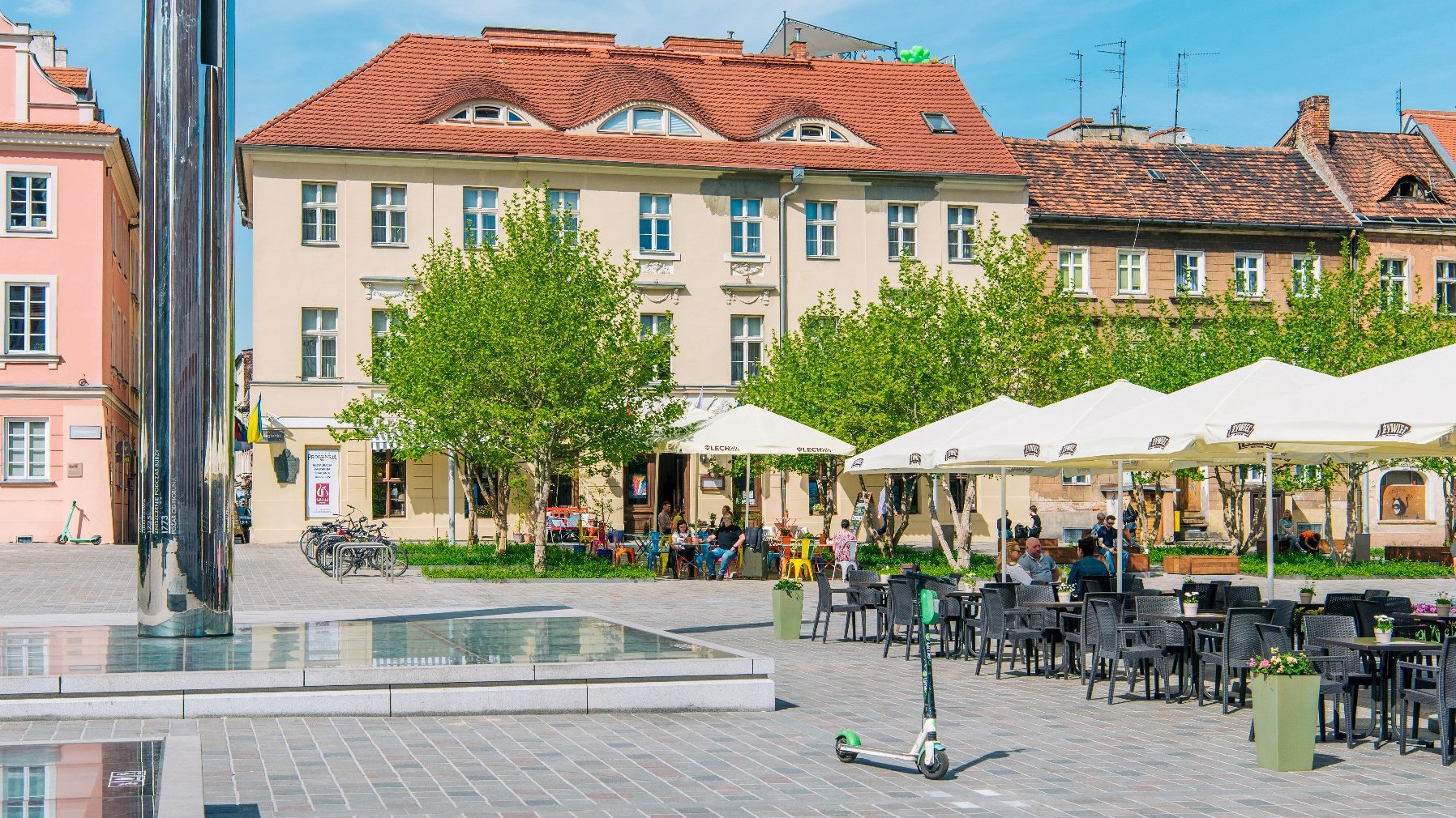 Na zdjęciu plac Kolegiacki, na pierwszym planie Zegar Historii, w tle drzewa i ogródek kawiarniany