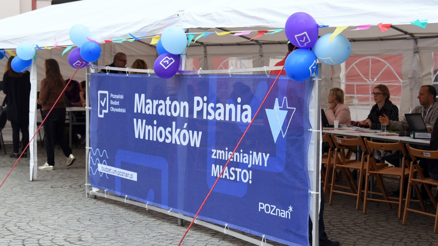 Zdjęcie przedstawia plakat z napisem "Maraton pisania wniosków". Z nim znajdują się długie stoły, przy których siedzą ludzie.