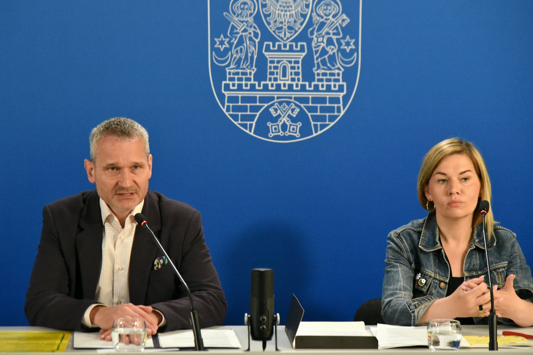 Na zdjęciu mężczyzna i kobieta za stołem konferencyjnym, za nimi gratatowa ścianka z herbem miasta - grafika rozmowy