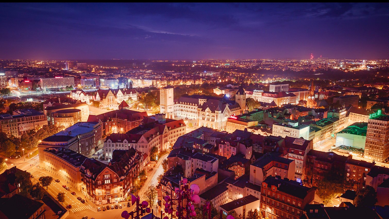 Na zdjęciu panorama Poznania po zmroku, w centrum zabudowa śródmiejska widziana z lotu ptaka - grafika rozmowy