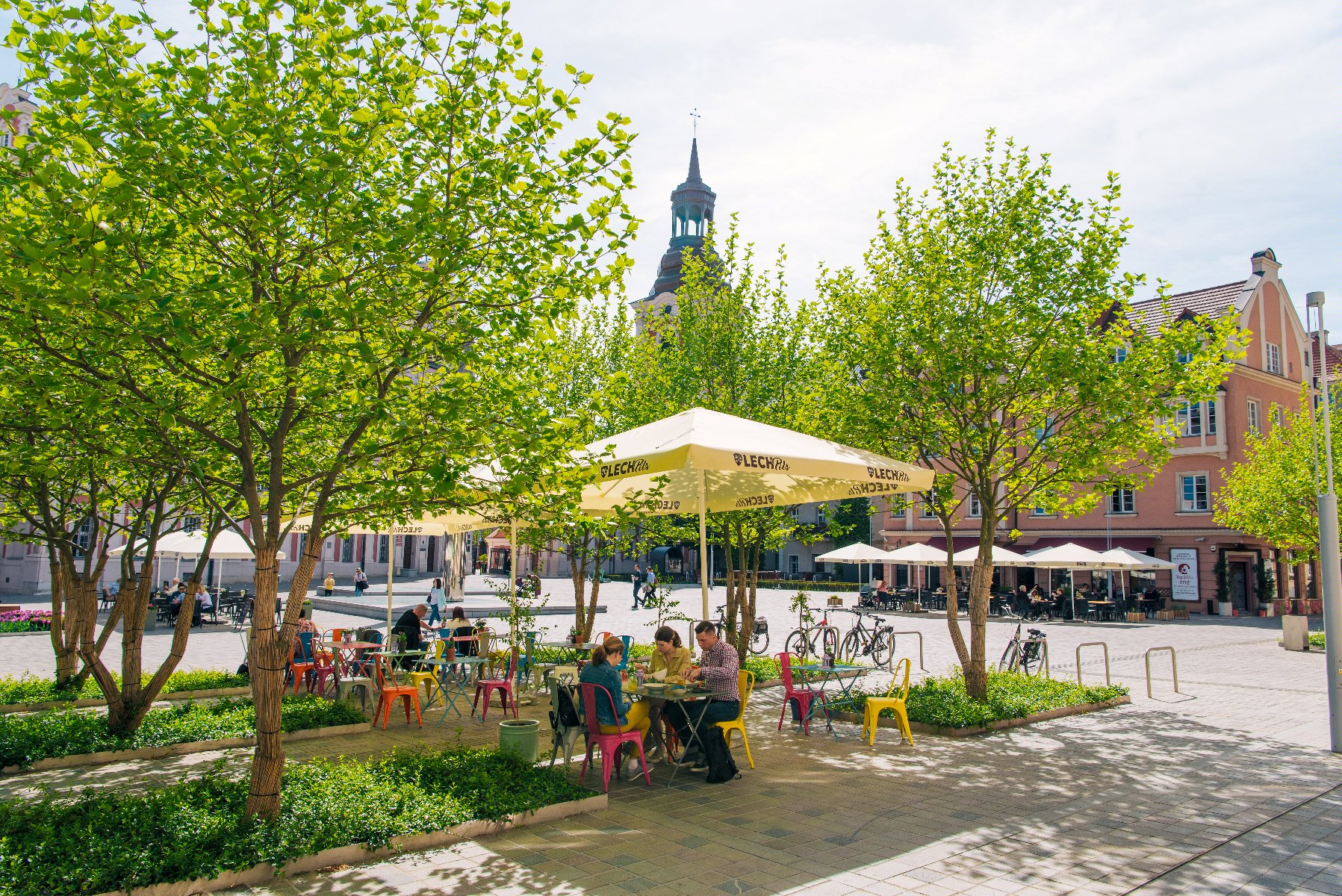 Na zdjęciu pl. Kolegiacki. Na pierwszym planie ogródek restauracyjny otoczony drzewami. W tle wieża poznańskiej Fary i kamienice. - grafika rozmowy