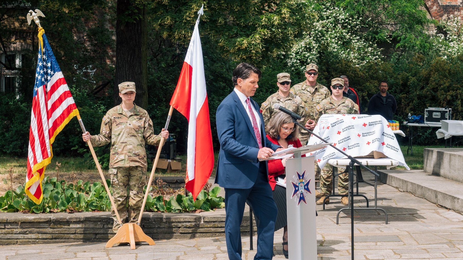 Na zdjęciu mężczyzna przy mikrofonie, w tle żołnierze i dwie flagi - polska i amerykańska