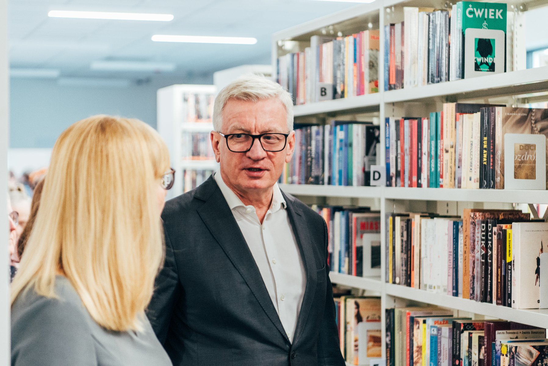 Na zdjęciu prezydent Poznania rozmawia z dyrektorką biblioteki (tyłem), w tle regał z książkami - grafika rozmowy