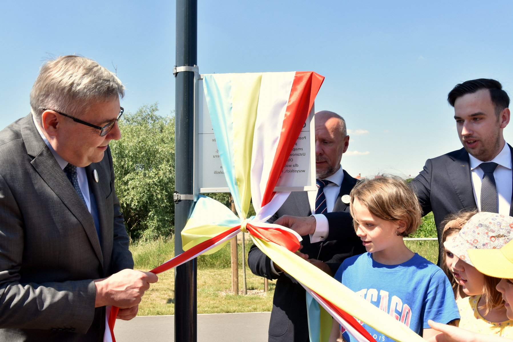 Na zdjęciu kilka osób - trzej dorośli mężczyźni oraz kilkoro dzieci - odsłaniają tabliczkę z nazwą skweru, ciągnąc za wstęgi w kolorach flag - grafika rozmowy