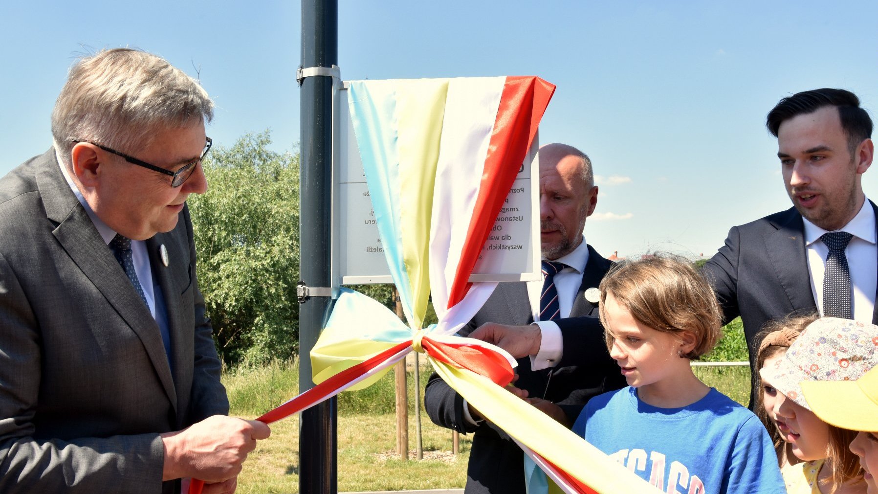 Na zdjęciu kilka osób - trzej dorośli mężczyźni oraz kilkoro dzieci - odsłaniają tabliczkę z nazwą skweru, ciągnąc za wstęgi w kolorach flag