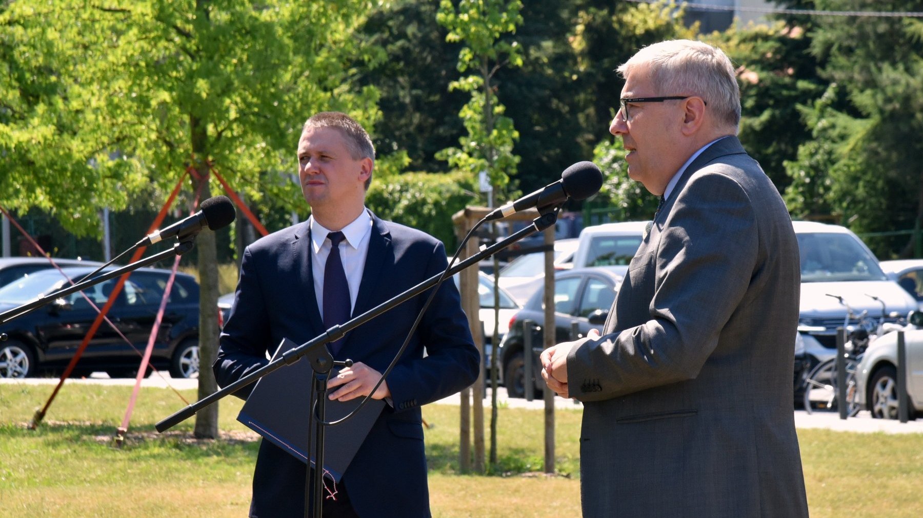 Na zdjęciu dwóch mężczyzn przy mikrofonach: przewodniczący rady miasta i dyrektor Gabinetu Prezydenta. W tle drzewa i samochody