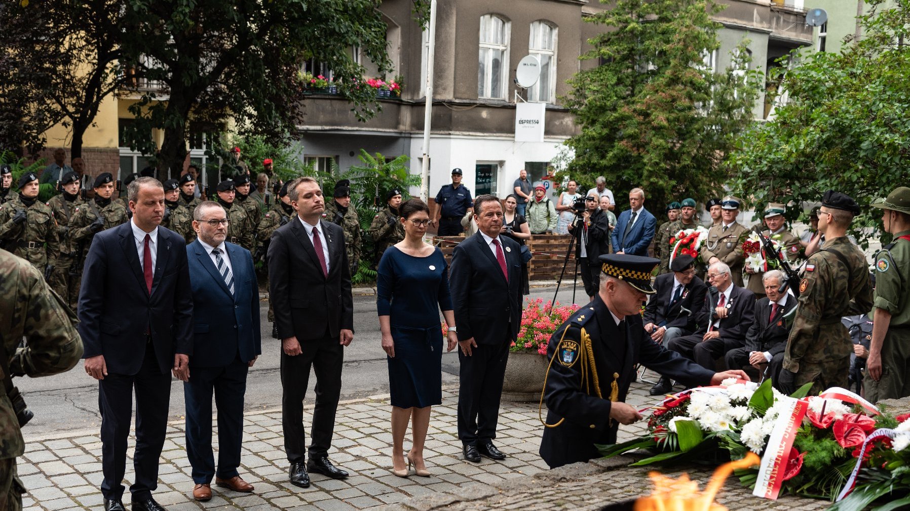 Na zdjęciu delegacja składająca kwiaty, widać strażnika miejskiego z wiązanką, na pierwszym planie płonący znicz