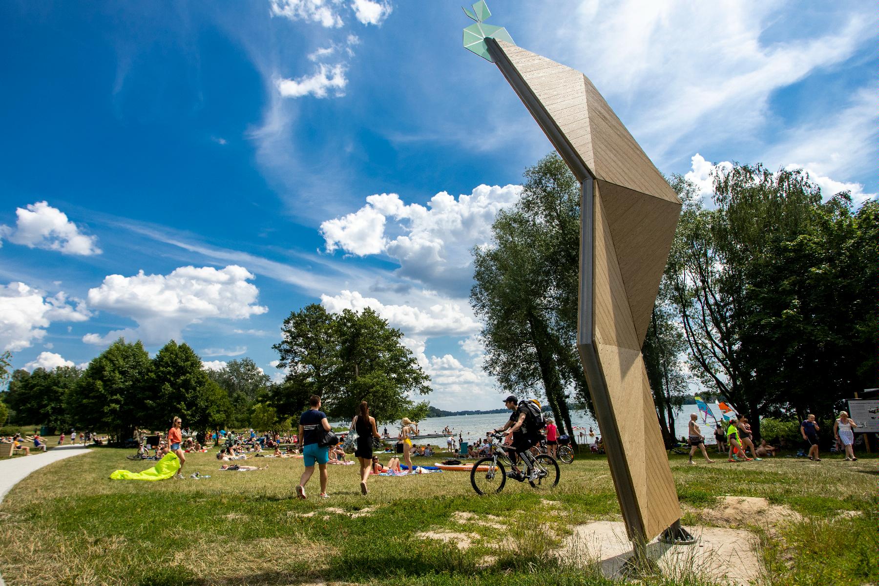 Zdjęcie przedstawia plażę nad jeziorem Kierskim. Widać na nim ludzi opalających się, spacerujących i jeżdżących na rowerach. W tle znajduje się woda. - grafika rozmowy