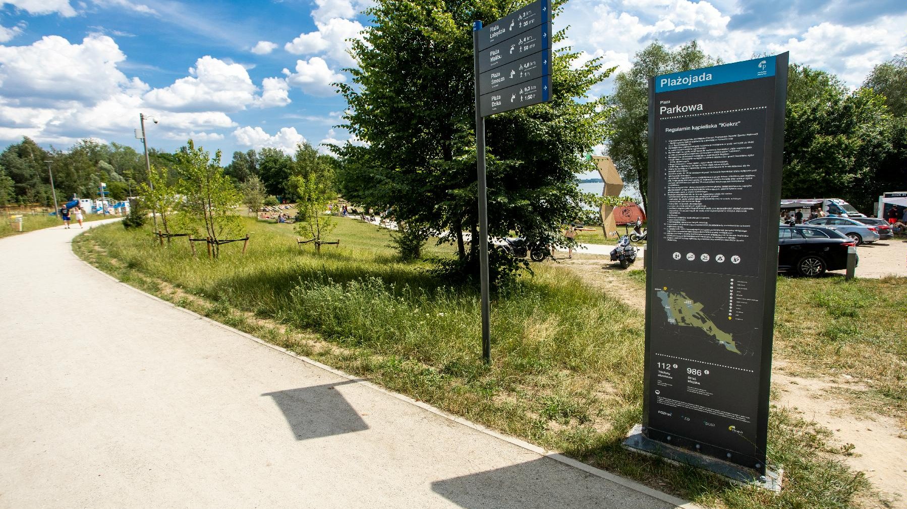 Zdjęcie przedstawia trawiastą plażę oraz tablice. Jedna to drogowskaz, druga informauje o zasadach panujących na plaży.