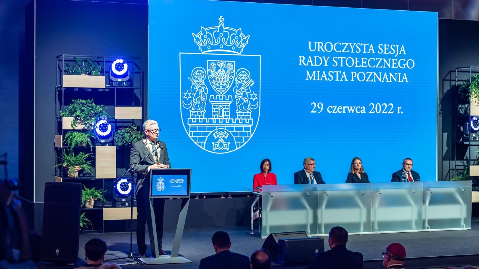 Na zdjęciu prezydent Poznania przy mównicy, obok przewodniczący i wiceprzewodniczący rady miasta, w tle ekran z herbem miasta