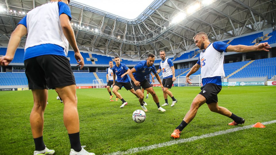 Grupa piłkarzy podaje piłkę miedzysobą. W tle stadion.