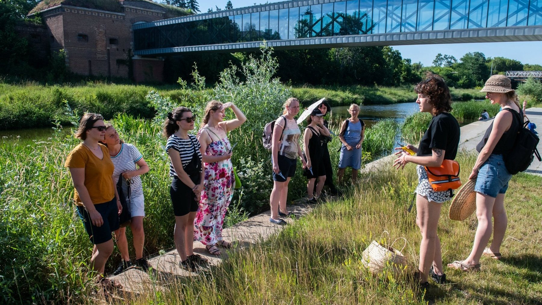 Na zdjęciu grupa ludzi stojących nad rzeką, dwie osoby tłumaczą coś pozostałym