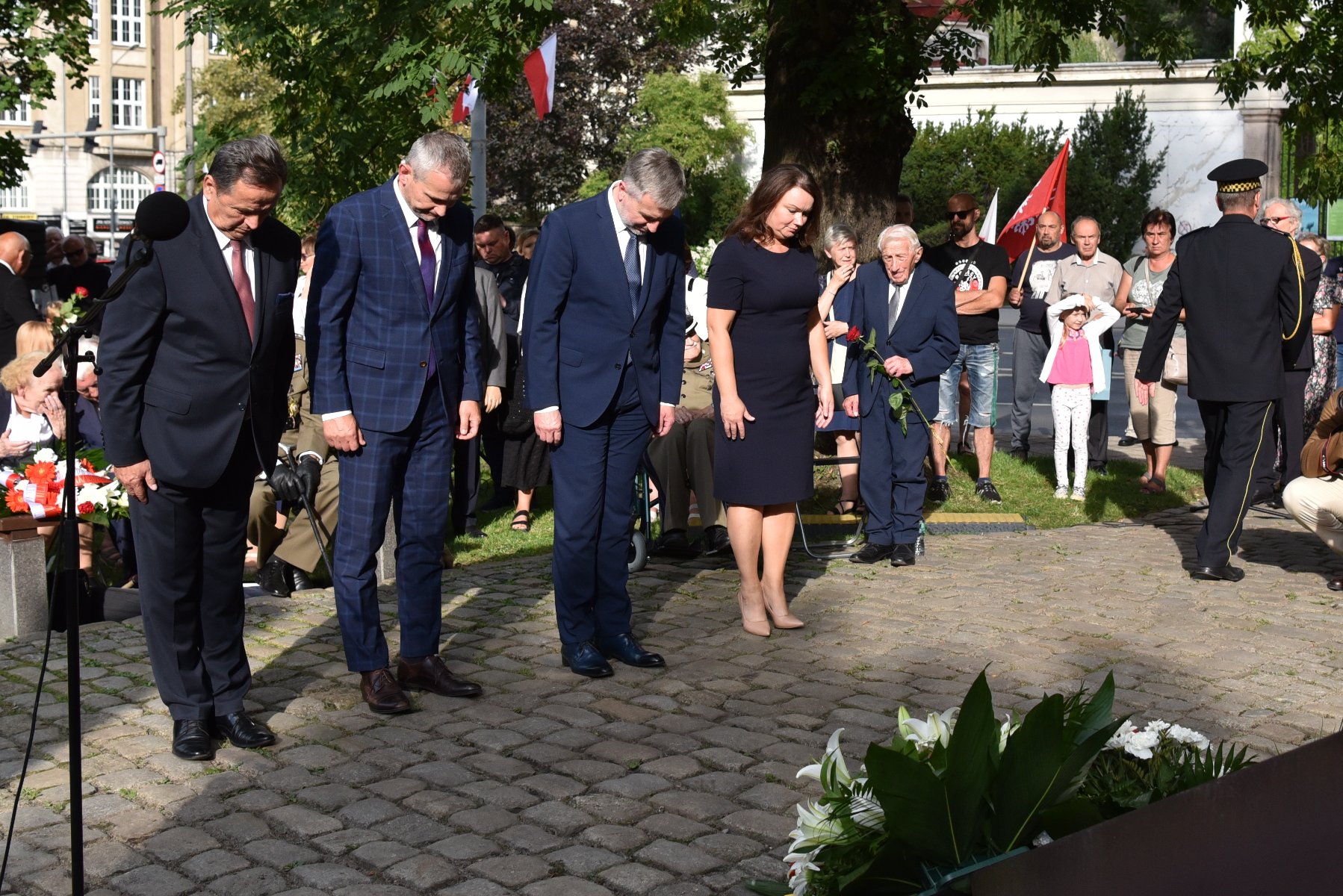 Na zdjęciu przedstawiciele władz rządu i samorządu składają wiązankę kwiatów, pochyleni - grafika rozmowy