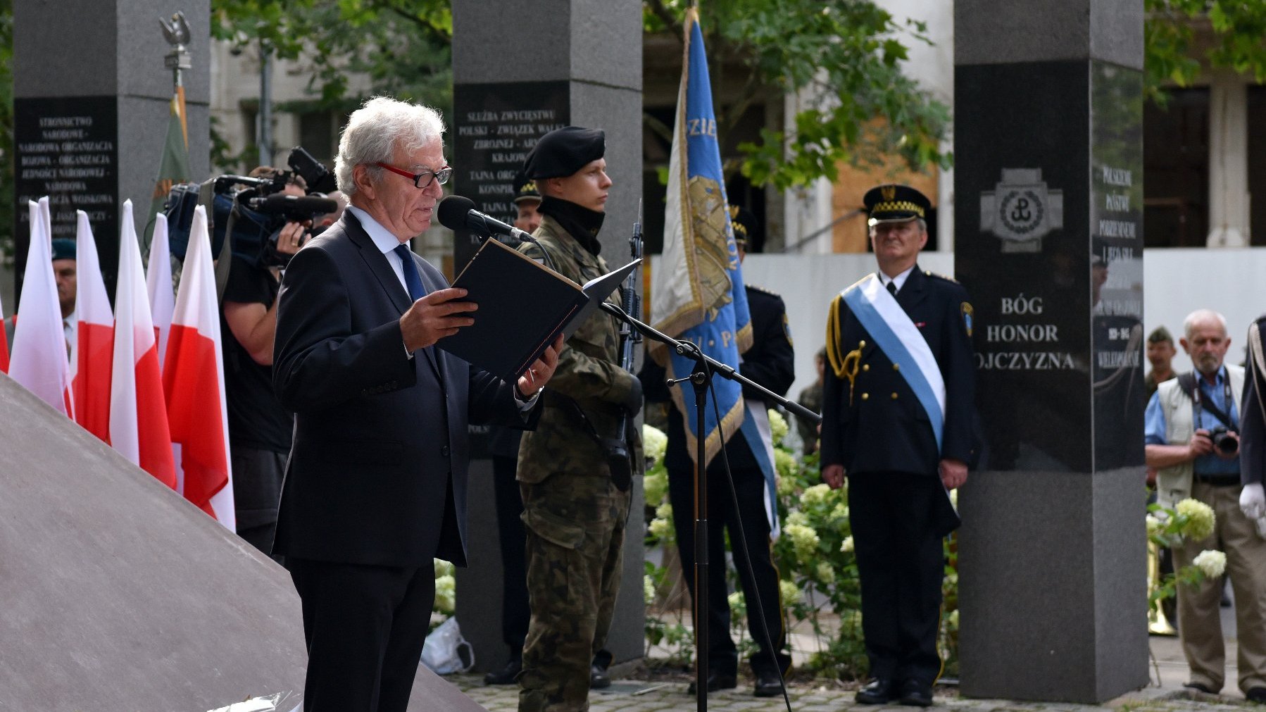 NA zdjęciu mężczyzna w garnitirze, przy mikrofonie, pod pomnikiem, w tle żołnierze