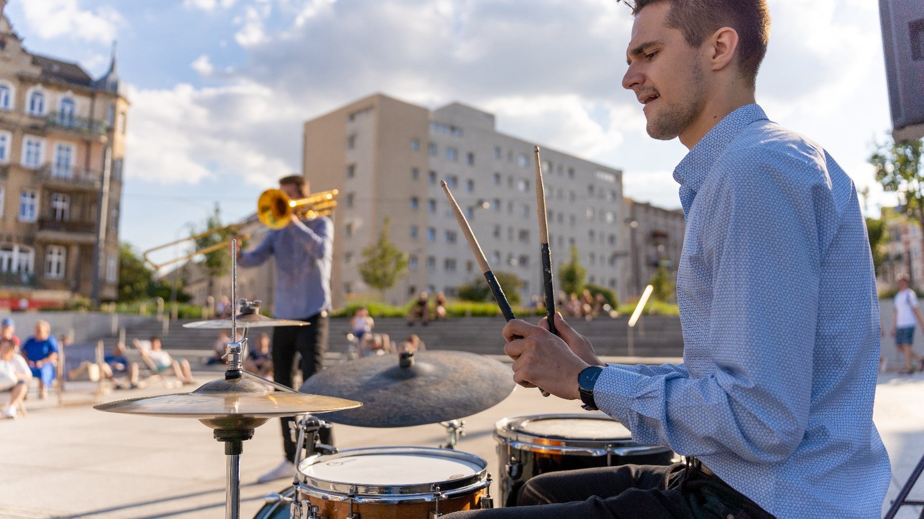 na zdjęciu mężczyzna grający na perkusji, w tle drugi, grający na instrumencie dętym, obok publiczność